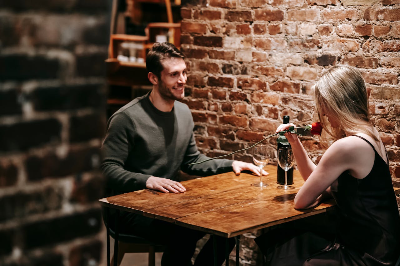 services-03 Young couple in elegant clothes sitting in light cafeteria at wooden table near brick wall and having romantic dinner with red rose and wine near glasses