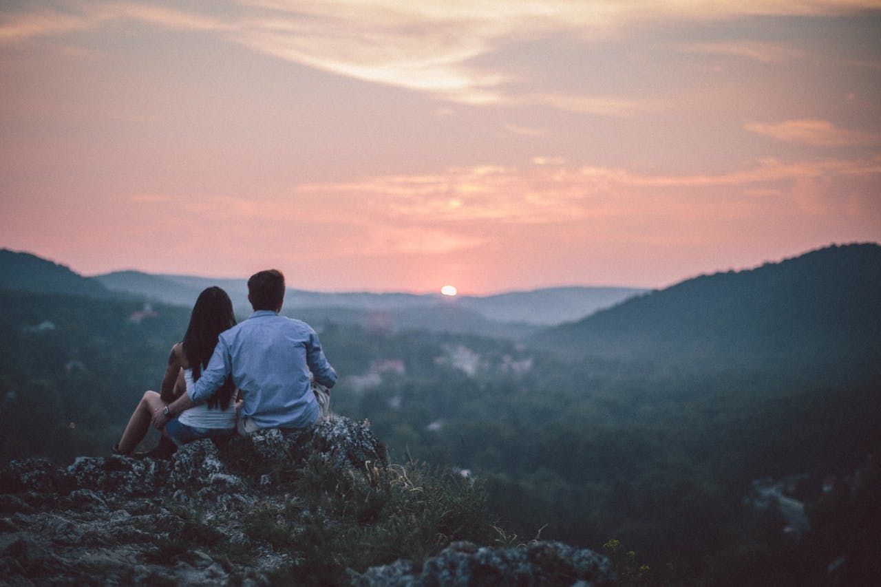 who-we-are A couple sits on a hill in Budapest, watching the sunset over distant hills, symbolizing love and togetherness.
